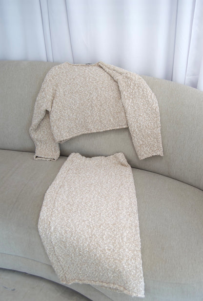 Picture features a two piece boucle material sweater and midi skirt set lying on a couch.