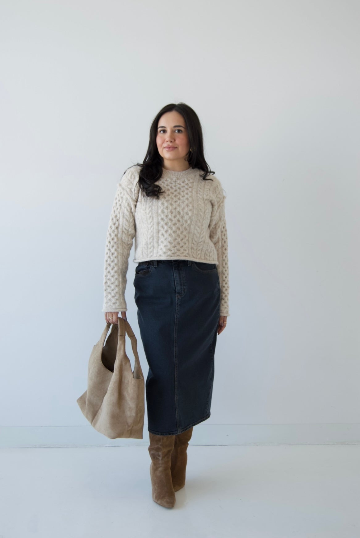 Picture features a neutral cable knit sweater on a model wearing a dark denim midi skirt, taupe suede boots and a taupe suede purse.