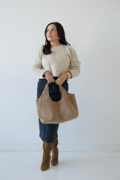 Picture features a neutral cable knit sweater on a model wearing a dark denim midi skirt, taupe suede boots and a taupe suede purse.