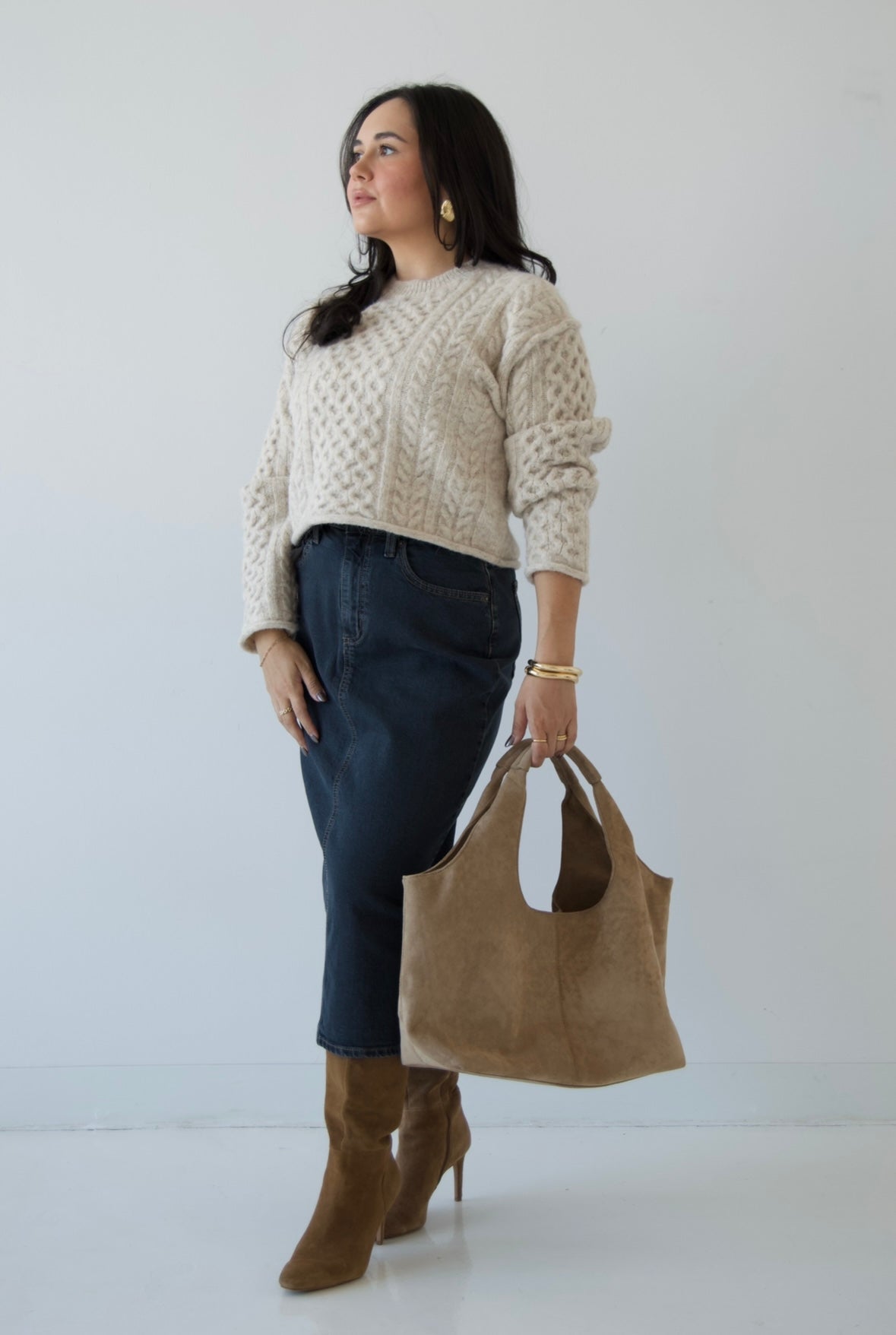 Picture features a neutral cable knit sweater on a model wearing a dark denim midi skirt, taupe suede boots and a taupe suede purse.