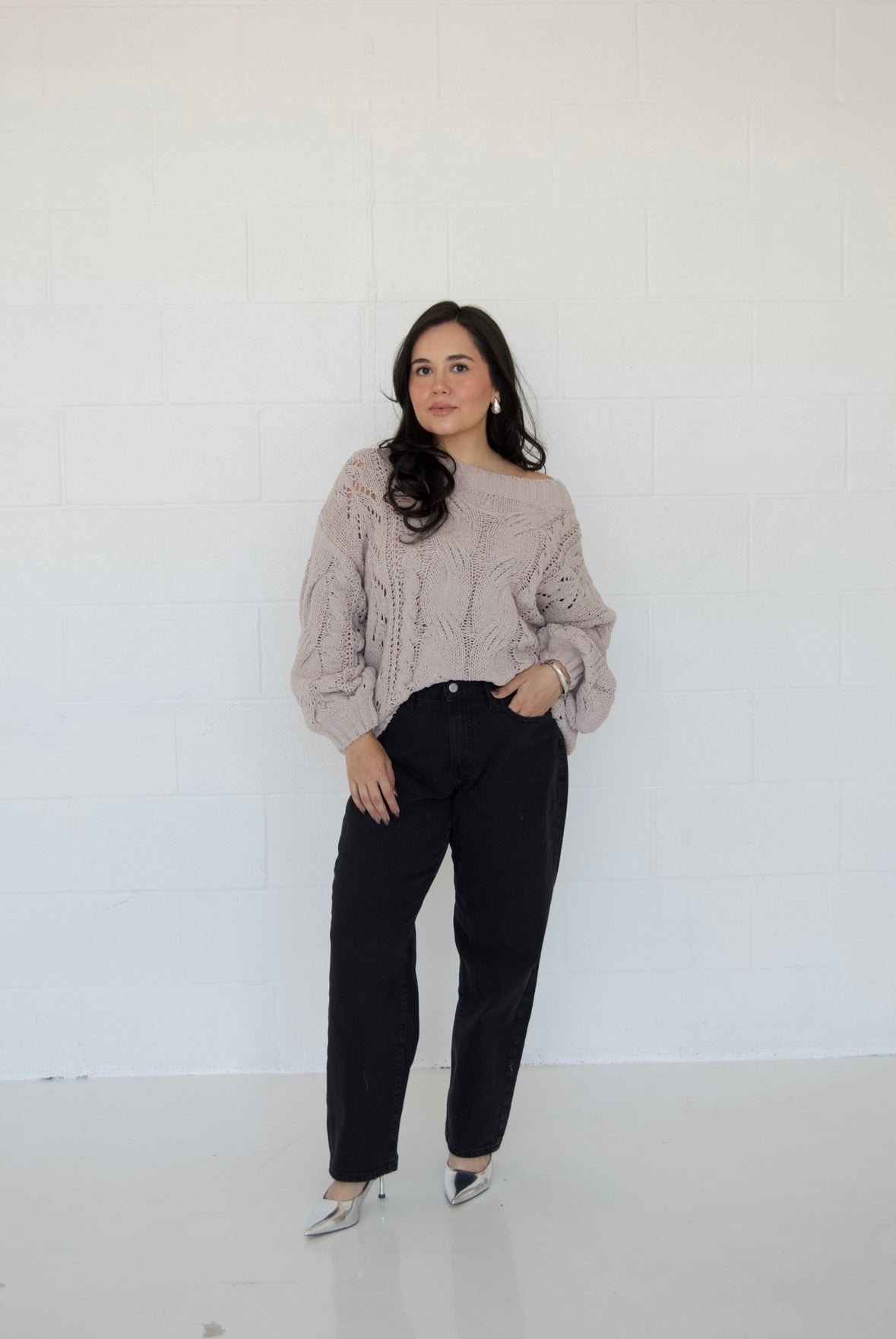 Picture features an off the shoulder neutral cable knit sweater on a model wearing dark jeans and silver heels.