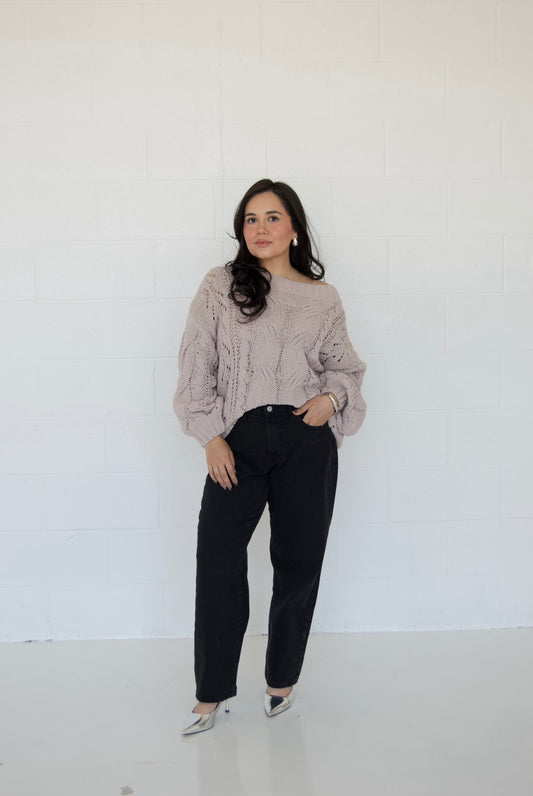 Picture features an off the shoulder neutral cable knit sweater on a model wearing dark jeans and silver heels.