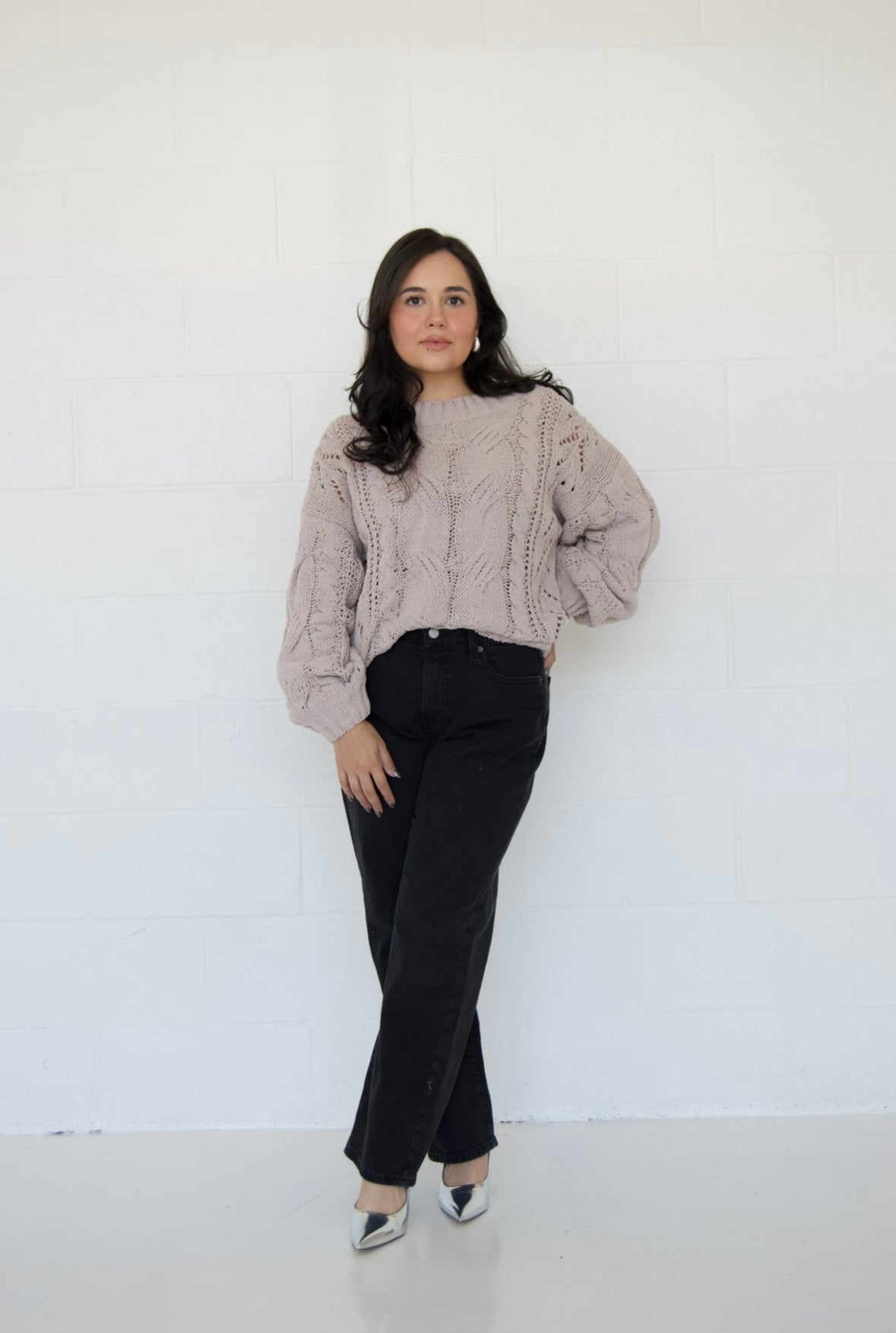 Picture features an off the shoulder neutral cable knit sweater on a model wearing dark jeans and silver heels.