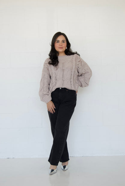 Picture features an off the shoulder neutral cable knit sweater on a model wearing dark jeans and silver heels.